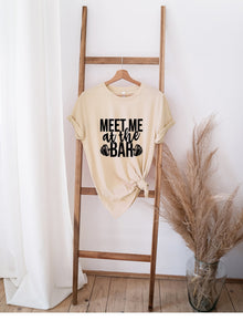 Beige "Meet Me at the Bar" t-shirt with black text, hanging on wooden ladder in minimalist room decor.