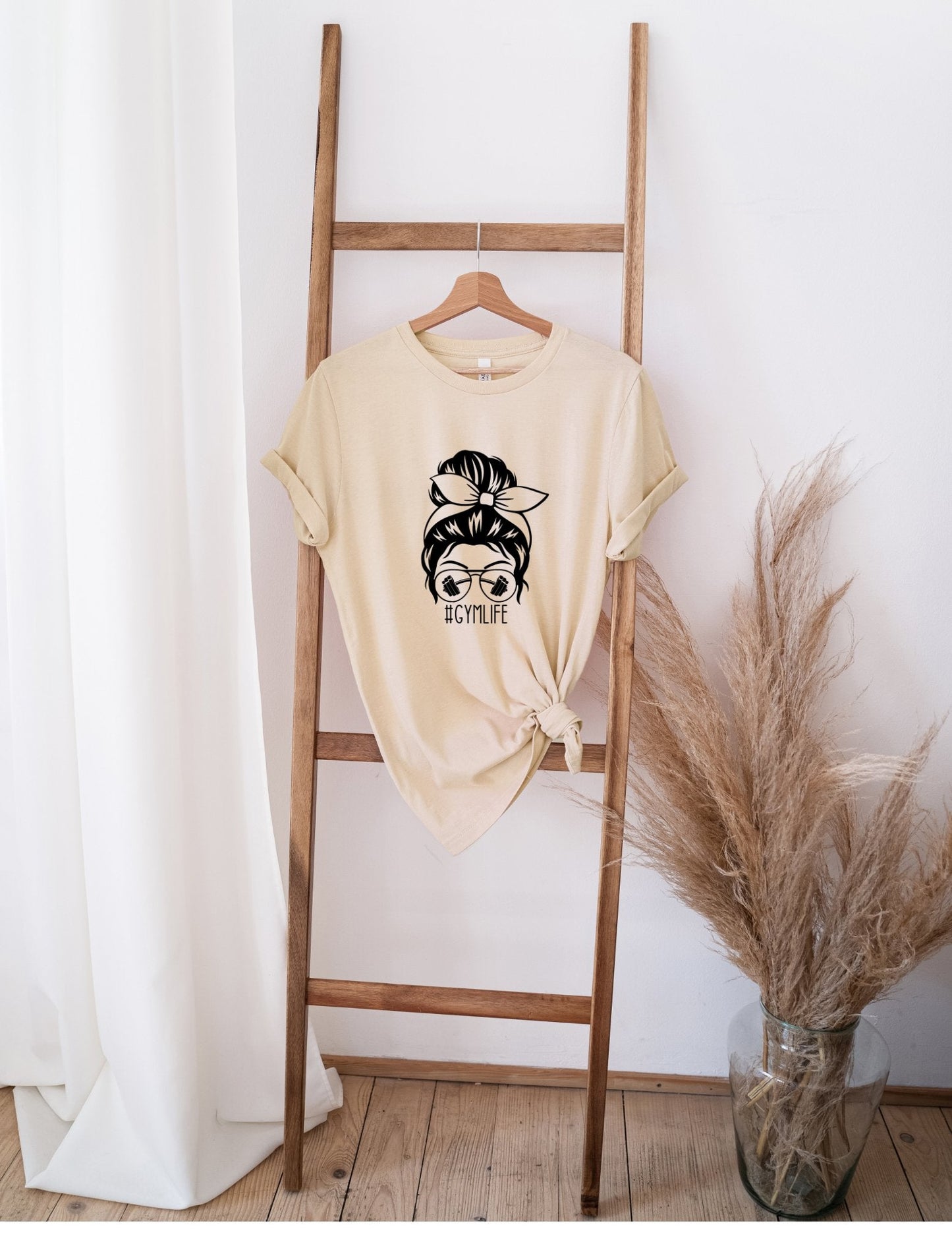 Beige Messy Bun GYM Life t-shirt with graphic print hanging on wooden ladder next to dried pampas grass.