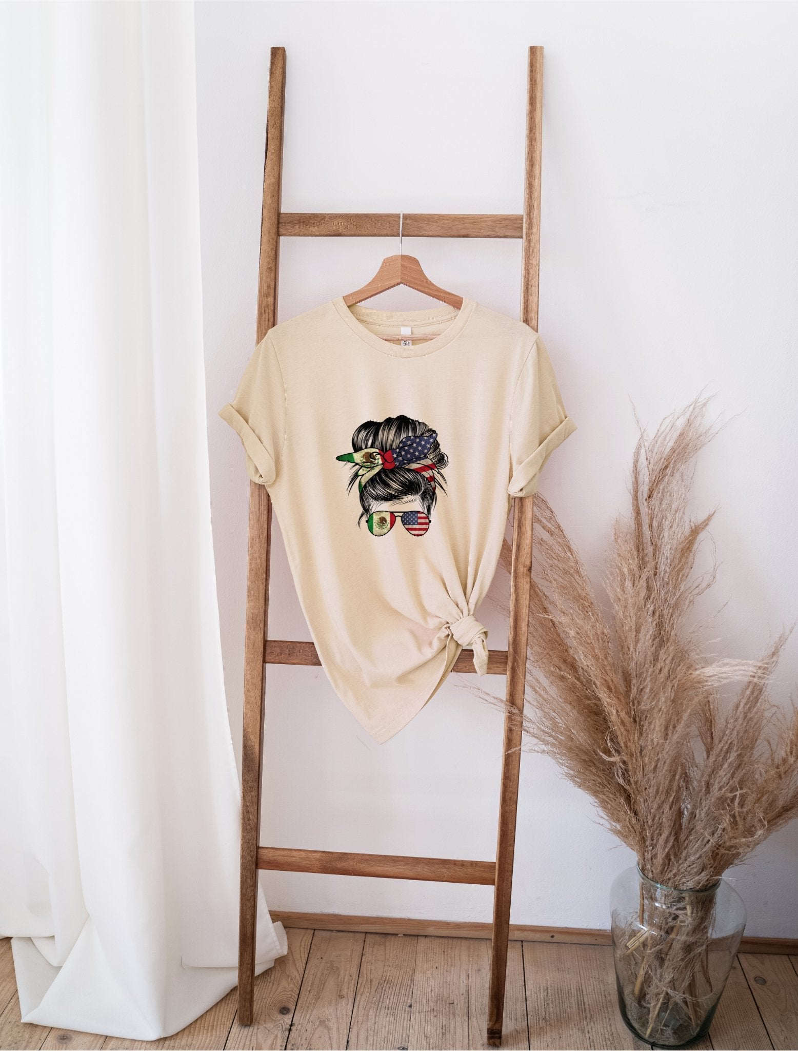 Mexamerican Messy Bun beige t-shirt with graphic print hanging on wooden ladder near pampas grass
