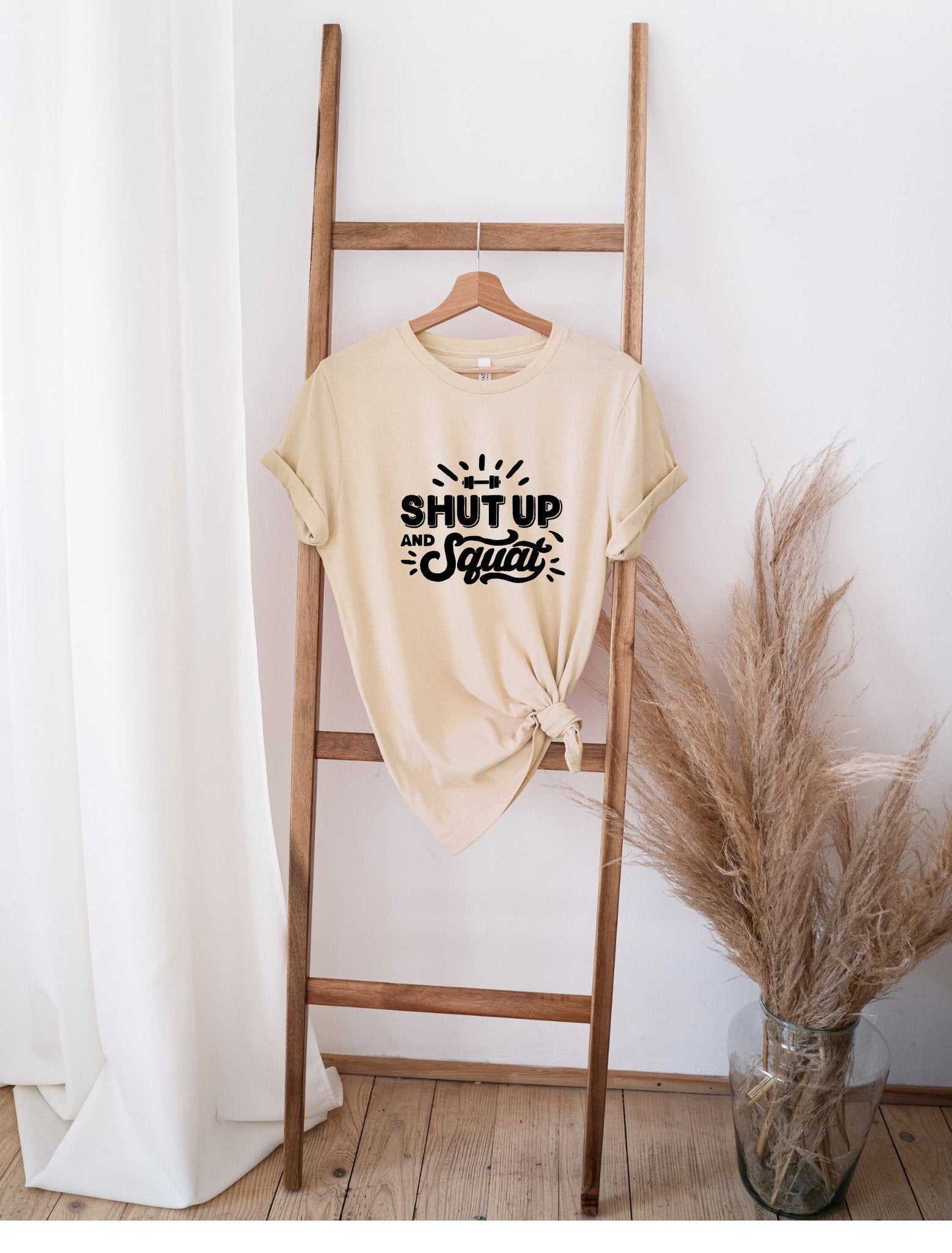 Beige "Shut Up and Squat" graphic t-shirt tied at the bottom hanging on wooden ladder against white wall with pampas grass decor