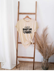 Beige "Shut Up and Squat" graphic t-shirt tied at the bottom hanging on wooden ladder against white wall with pampas grass decor