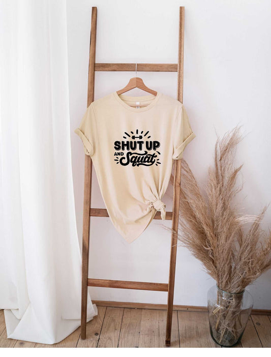 Beige "Shut Up and Squat" graphic t-shirt tied at the bottom hanging on wooden ladder against white wall with pampas grass decor