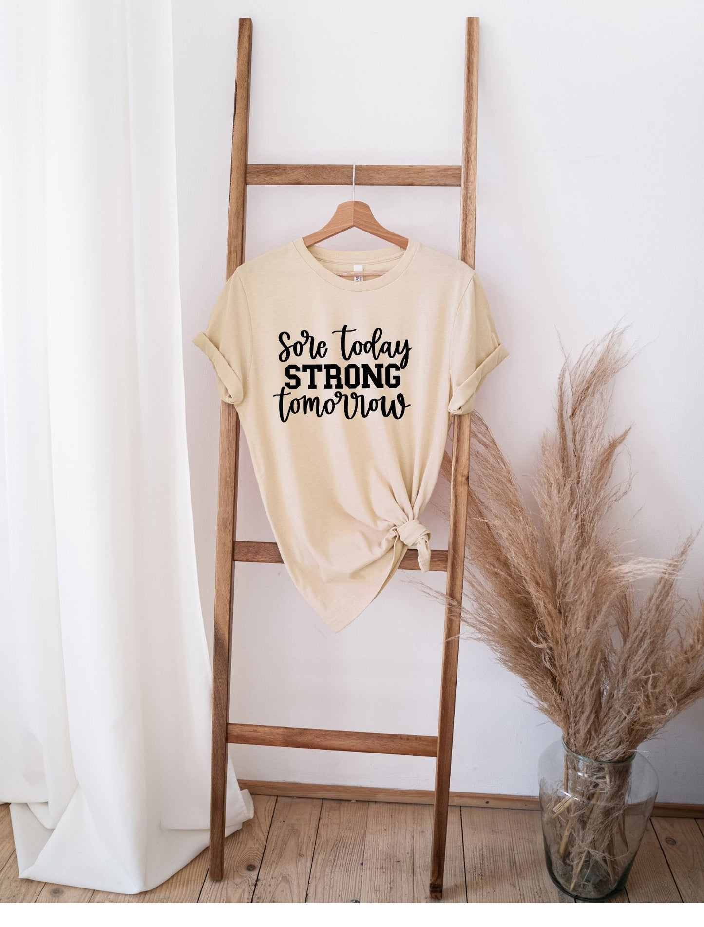 Beige Sore Today Strong Tomorrow t-shirt hanging on wooden ladder next to vase with dried pampas grass