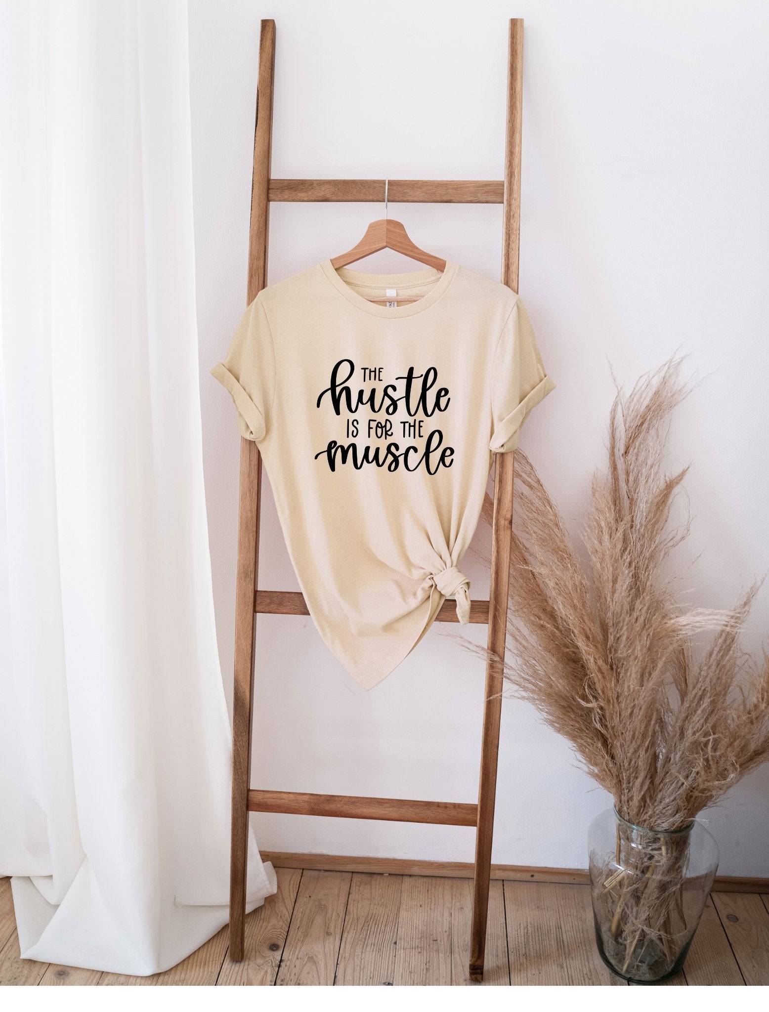 The Hustle for the Muscle beige t-shirt with black text hanging on wooden ladder next to dried pampas grass in vase